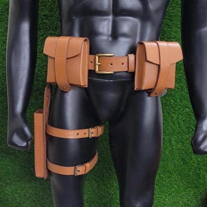 Peut inclure: Une ceinture utilitaire en cuir marron avec deux pochettes, une ceinture et une sangle de jambe. La ceinture a une boucle dorée. La sangle de jambe a deux sangles réglables avec des boucles dorées. Les pochettes sont rectangulaires avec une fermeture à rabat.
