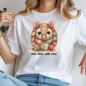 May include: White t-shirt featuring a colorful, patchwork-style armadillo illustration. The armadillo has large ears and a sweet expression. Below the image is the text "HARD SHELL, KIND SOUL."