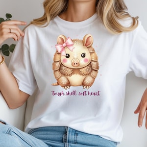 May include: White t-shirt featuring a watercolor illustration of a cute armadillo with a pink bow. The armadillo has a brown shell and is printed with the text "Tough shell, soft heart" in a purple script. The shirt is a casual style.
