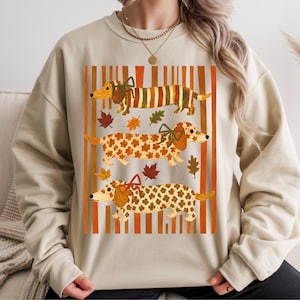 Puede incluir: Una sudadera beige de cuello redondo con un estampado gráfico de tres perros salchicha. Los perros están decorados con motivos otoñales, como rayas y hojas, y llevan pequeños lazos. El fondo presenta rayas verticales de color naranja.