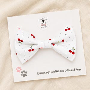 May include: A white fabric bowtie featuring a pattern of small red cherries and black dots, attached to a display card with the text Sweet Snout BOWTIES and Handmade bowties for cats and dogs, including a pink and gray paw print graphic.