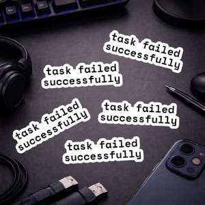May include: White stickers with the text "task failed successfully" in a black font. The stickers are scattered on a dark surface, along with a keyboard, headphones, a phone, and a pen.