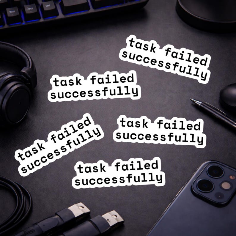 May include: White stickers with the text "task failed successfully" in a black font. The stickers are scattered on a dark surface, along with a keyboard, headphones, a phone, and a pen.