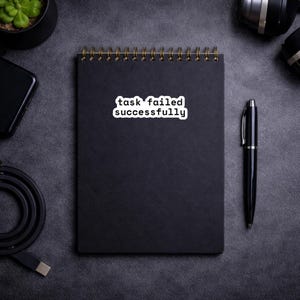 May include: A black spiral-bound notebook with the text "task failed successfully" on a white sticker. A black pen rests beside the notebook. Other items include a smartphone, a small potted plant, and camera equipment, all on a dark gray surface.