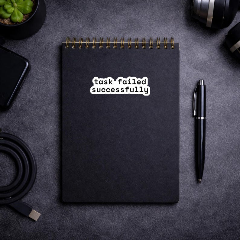 May include: A black spiral-bound notebook with the text "task failed successfully" on a white sticker. A black pen rests beside the notebook. Other items include a smartphone, a small potted plant, and camera equipment, all on a dark gray surface.