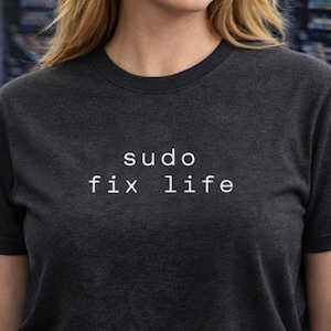 Può includere: T-shirt grigio scuro con la scritta bianca "sudo fix life". Il testo è centrato sul petto. La maglietta è a girocollo.