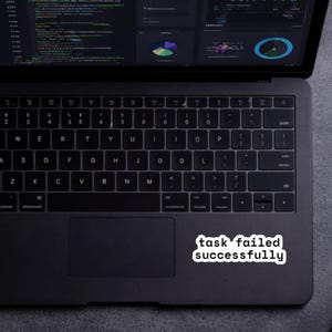 May include: A black laptop with a keyboard and a white sticker that reads "task failed successfully." The laptop screen displays code and data visualizations. The laptop is on a gray surface.