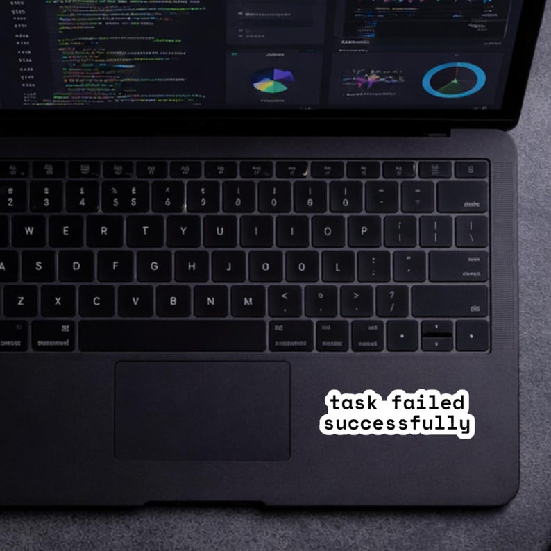 May include: A black laptop with a keyboard and a white sticker that reads "task failed successfully." The laptop screen displays code and data visualizations. The laptop is on a gray surface.