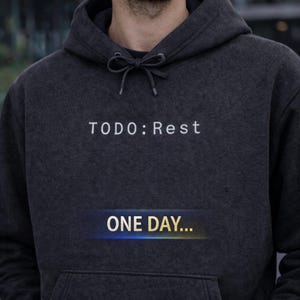 May include: Dark gray hoodie with the text "TODO: Rest" in white above a blue and yellow gradient banner that reads "ONE DAY...". The hoodie has a drawstring hood and a front pocket.
