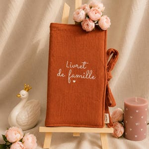 Puede incluir: Una funda de tela color óxido para libro de familia con el texto bordado en blanco "Livret de famille" y un pequeño icono de corazón. La funda incluye un cierre de lazo de tela a juego y se muestra sobre un pequeño caballete de madera.