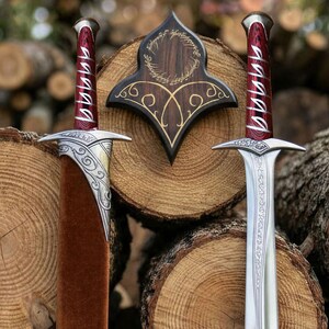 Könnte beinhalten: Zwei Schwerter mit Griffen aus rotem Maserholz, verziert mit silberner Elbenschrift, silbernen Parierstangen mit filigranen Verzierungen und einer braunen Samtscheide. Eine dunkle Wandplakette aus Holz zeigt eine goldene Inschrift.