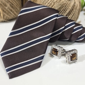 May include: A brown necktie featuring diagonal white and navy blue stripes, paired with a set of square silver cufflinks. Each cufflink has a central brown stone surrounded by a border of small clear crystals in a polished metal setting.