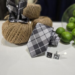 May include: A grey and white plaid tie and matching pocket square are displayed with a pair of square cufflinks. The accessories are on a white surface, with a background of twine and green limes.