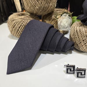 May include: A dark charcoal gray necktie with a textured fabric finish, folded neatly, accompanied by a pair of square silver-toned cufflinks featuring a black geometric Greek key pattern design on the front face of each piece.