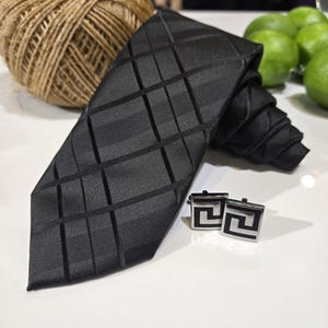 May include: A black tie with a geometric design and matching square cufflinks. The tie is folded, and the cufflinks feature a black and silver pattern. A ball of twine and limes are in the background.