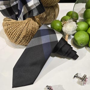May include: A black tie with a blue and grey check pattern, a matching pocket square, and silver cufflinks with pink square stones. The items are arranged on a white surface with green limes and twine in the background.