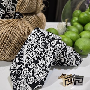 Op de afbeelding: Een zwart-witte paisley stropdas en bijpassende manchetknopen. De manchetknopen hebben een goud-zwart geometrisch ontwerp. De stropdas ligt op een wit oppervlak, met touw en limoenen op de achtergrond.