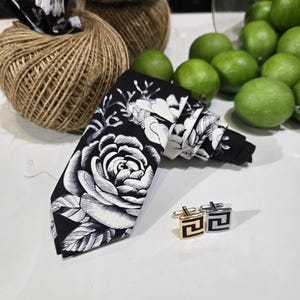 May include: A black tie with a white floral design, alongside gold and silver cufflinks. The tie showcases a large rose motif. Jute twine and green limes are in the background.