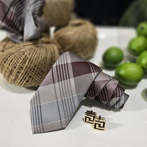 Puede incluir: Una corbata a cuadros en tonos grises y burdeos, junto con gemelos dorados con un diseño geométrico negro. La corbata está dispuesta sobre una superficie blanca, lo que sugiere un entorno formal o de negocios.