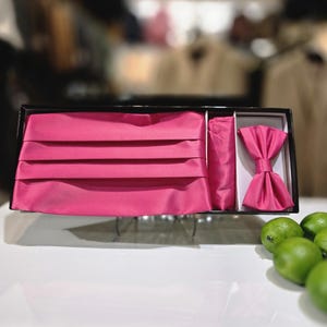 May include: A formalwear set in a black box, featuring a bright pink cummerbund, bow tie, and pocket square. The cummerbund is pleated, and the bow tie is pre-tied. The set is displayed on a white surface with green limes.
