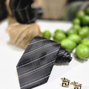Peut inclure: Une cravate noire à rayures blanches et beiges, enroulée, à côté de boutons de manchette dorés avec des motifs géométriques noirs. La cravate est sur une surface blanche, avec des fruits verts flous en arrière-plan.