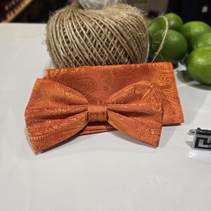 Peut inclure: Un ensemble de nœud papillon et pochette orange avec un motif cachemire. Le nœud papillon est pré-noué et de forme classique. La pochette est pliée. Un bouton de manchette argenté est visible. L'ensemble est sur une surface blanche.