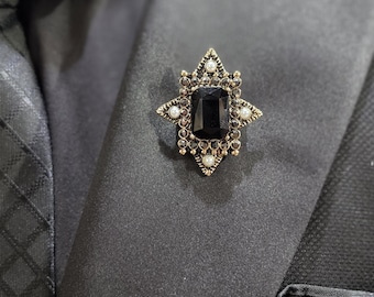 May include: A star-shaped brooch featuring a large rectangular black centre stone surrounded by smaller dark crystals and four small white pearls. The setting is an antique gold tone metal, pinned to the lapel of a black textured suit jacket.