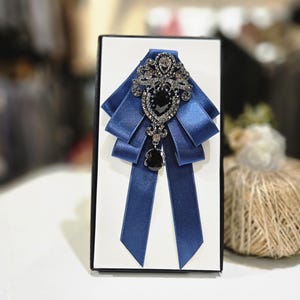 May include: A blue satin bow tie with a decorative brooch. The brooch features a large black gem surrounded by smaller clear gems. The bow tie is presented on a white background inside a black box.