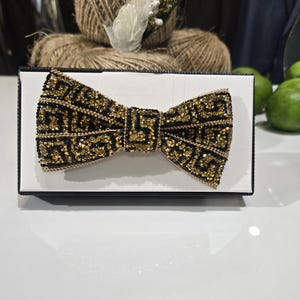 May include: A black and gold bow tie with a geometric pattern, presented on a white surface inside a black box. The bow tie is heavily embellished with gold sequins and black accents, creating a sparkling effect.