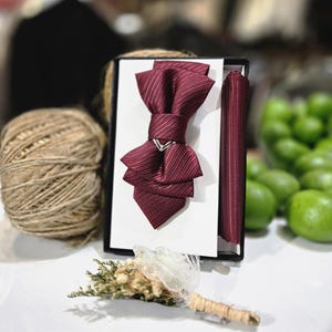 Puede incluir: Un conjunto de pajarita y pañuelo de bolsillo burdeos con un patrón de rayas, presentado en una caja negra. También se ve un pequeño ojal con flores secas y una bola de hilo. El conjunto es probablemente para vestir formal.
