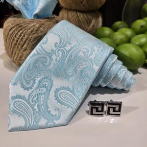 May include: A light blue paisley tie and pocket square are displayed with black and silver cufflinks. The tie is rolled up, showing the intricate pattern. The items are part of a formal wear set. The tie and pocket square are made of a silky material.