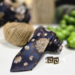 Puede incluir: Una corbata azul marino con un estampado floral en dorado y blanco, combinada con gemelos cuadrados dorados y negros. La corbata está enrollada, mostrando el intrincado diseño. Una bola de hilo y objetos verdes están en el fondo.