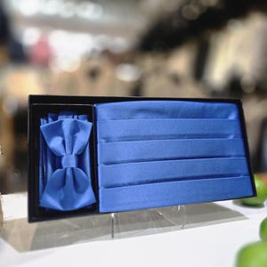 May include: A formal wear set in a black box, featuring a vibrant blue bow tie and cummerbund. The bow tie is pre-tied, and the cummerbund has horizontal pleats. The set is displayed on a white surface with a blurred background.