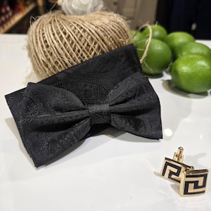 May include: A black paisley bow tie and gold cufflinks with a black Greek key design are displayed on a white surface. A ball of twine and green limes are in the background, suggesting a theme of formal wear accessories.