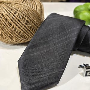 May include: A dark grey plaid tie with a classic design. The tie is made of a woven fabric and features a subtle grid pattern. A silver cufflink is visible in the background. Suitable for formal or business wear.