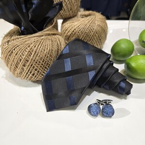 Peut inclure: Une cravate noire et bleue à motifs et des boutons de manchette assortis sont présentés sur une surface blanche. La cravate a un motif à carreaux avec des rayures bleues. Les boutons de manchette sont ronds avec une finition bleue scintillante.