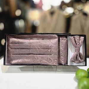 Puede incluir: Un conjunto de ropa formal en una caja negra, con un fajín, pajarita y pañuelo de bolsillo con estampado de cachemira en color malva. El conjunto se exhibe sobre una superficie blanca.