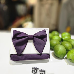 May include: A formal set featuring a deep purple bow tie, pocket square, and cufflinks. The bow tie and pocket square are displayed in a white box. The cufflinks are silver and black. Green limes are in the background.