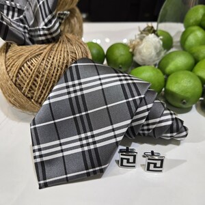 Peut inclure: Un ensemble cravate, pochette et boutons de manchette à carreaux gris, noirs et blancs. La cravate et la pochette sont pliées et disposées avec des boutons de manchette carrés avec un motif géométrique noir. L'ensemble est présenté avec une pelote de ficelle et des citrons verts.