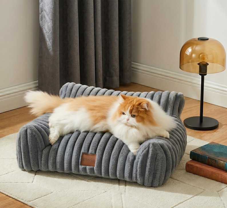 May include: A grey, corduroy-style pet sofa with a rectangular shape and a small label. An orange and white cat is lying on the sofa. The sofa has a curved back and armrests.
