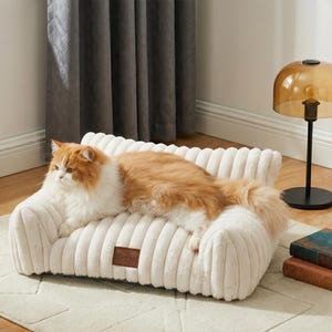 May include: A cream-coloured pet bed with a corduroy texture, resembling a miniature sofa. An orange and white cat is lounging on the bed. A small brown leather tag with the word "Sterish" is on the front of the bed.