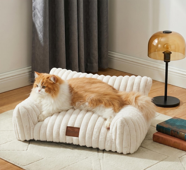May include: A cream-coloured pet bed with a corduroy texture, resembling a miniature sofa. An orange and white cat is lounging on the bed. A small brown leather tag with the word "Sterish" is on the front of the bed.