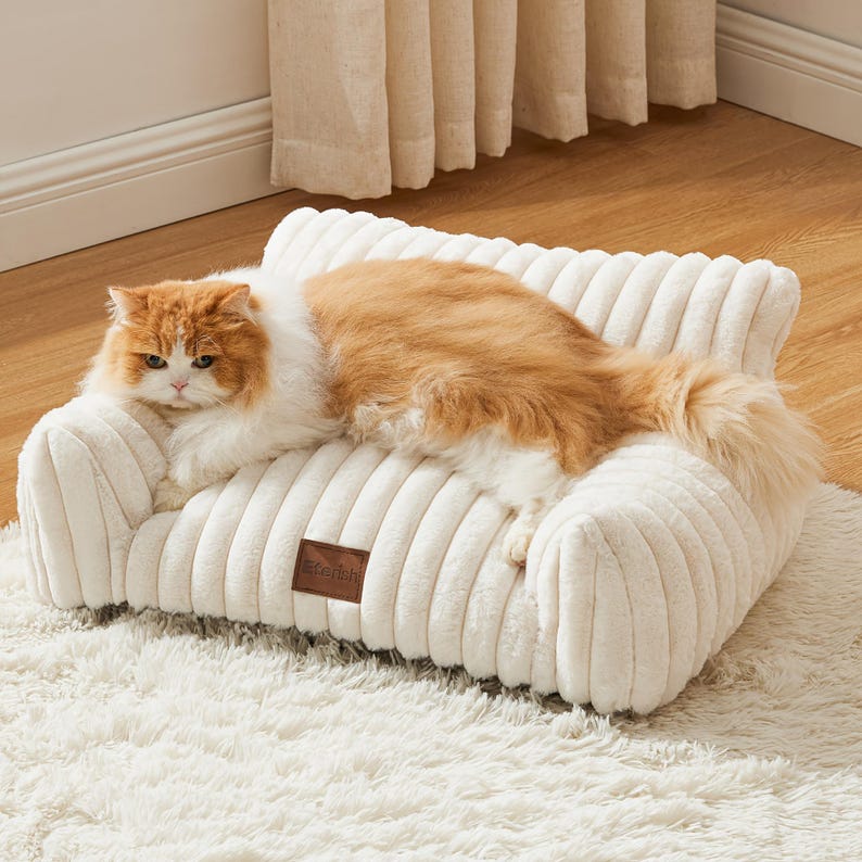 May include: A cream corduroy pet sofa with a brown tag. A ginger and white cat is resting on the sofa. The sofa is on a white, shaggy rug. The sofa is designed for a pet.