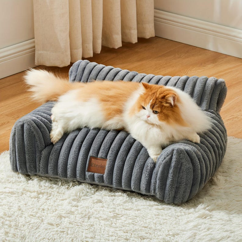 May include: A grey, corduroy-style pet sofa with a fluffy, orange and white cat resting on it. The sofa has a small, rectangular tag with text. The cat is lying down on a white, fluffy rug.
