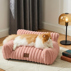 May include: A pink corduroy cat sofa with a brown tag. A ginger and white cat is resting on the sofa. A black lamp with a gold shade is in the background. The sofa is approximately 60 cm long.