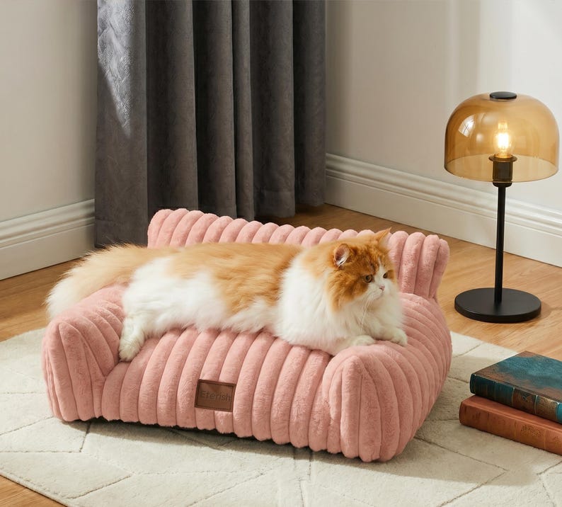 May include: A pink corduroy cat sofa with a brown tag. A ginger and white cat is resting on the sofa. A black lamp with a gold shade is in the background. The sofa is approximately 60 cm long.