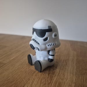 Può includere: Statuetta bianca e nera di un soldato delle tempeste di Star Wars. La statuetta è seduta con le gambe distese e le braccia lungo i fianchi. L'elmo del soldato delle tempeste ha copriocchi neri e un boccaglio nero. La statuetta è su una superficie di legno.