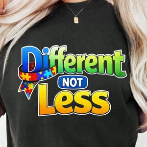 Könnte beinhalten: Schwarzes T-Shirt mit dem Schriftzug "Different NOT Less" in einem farbenfrohen Design. Das Wort "Different" ist in Blau und Grün gehalten, mit einem Band aus Puzzleteilen. Das Wort "Less" ist gelb.