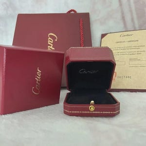 May include: A red Cartier ring box, open to reveal a black interior, sits next to a matching box and a document. The Cartier logo is visible on the boxes and the document. A red gift bag with a red cord is in the background.