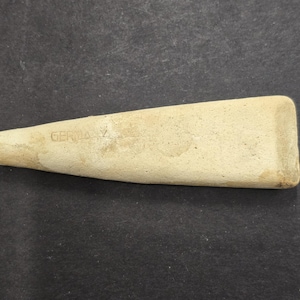 Puede incluir: Objeto en forma de cuña de color beige claro, con un extremo rectangular redondeado y una punta. La palabra "GERMANY" es débilmente visible en el lateral. El objeto parece estar hecho de un material similar a la piedra.